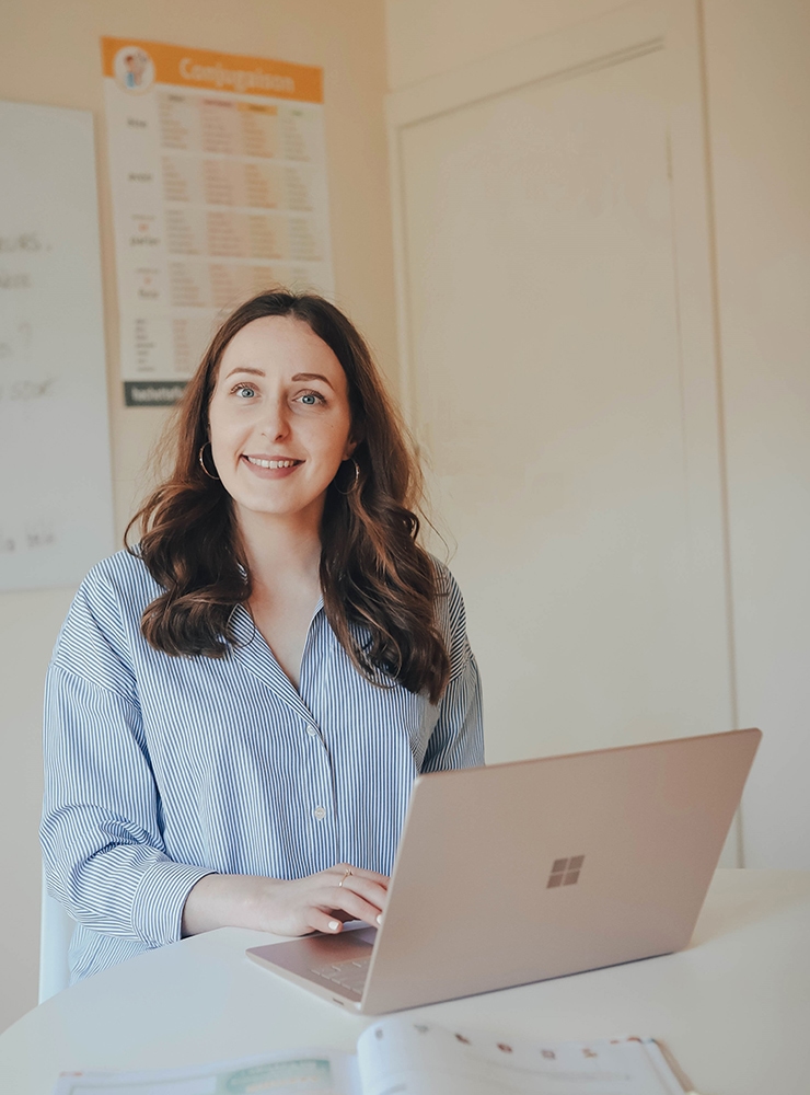 French Lessons For Beginners - Adelaide, French teacher with a welcoming smile, sitting at a desk with a laptop in a classroom, ready to begin a lesson for beginners with conjugation charts in the background., french for beginners,