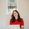 French classes, French teacher in a red shirt smiling at her laptop with a Paris poster in the background, ready to start an online French class.