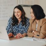 , Two women reviewing Advanced French lessons on a laptop in a classroom with French conjugation charts in the background., conversational french course, best tutoring near me, french language teacher, french centre, french speaking school, tutor french, french private tutor, learn french speaking, learn french course, french language centre,